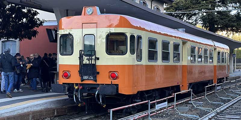 A restored vintage Italian train travelling towards Lake Como, with period carriages, mountains and the lake landscape beyond.